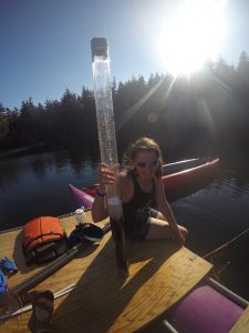 Caitlin holds a clear plastic tube of sediment with sunlight and pond in background