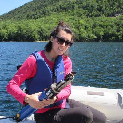 Rachel Fowler holding a water quality sensor on a boat with water and mountain in background