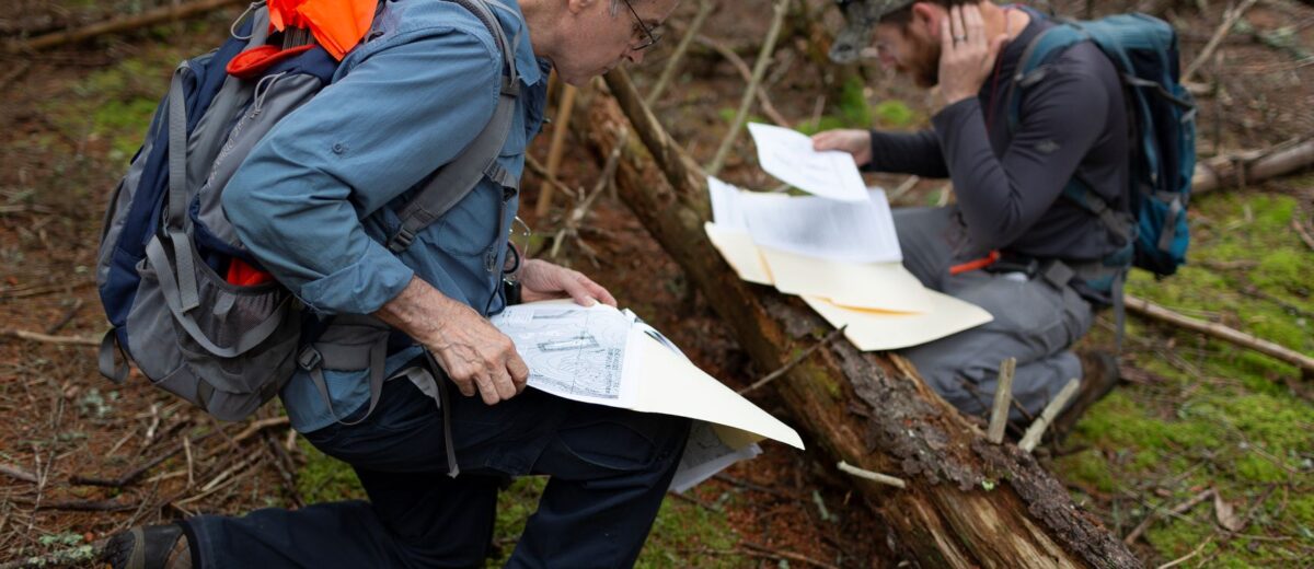 Two people wearing backpacks pause at a log in the woods to review maps